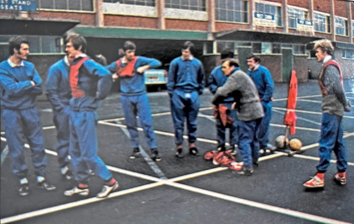 A group of Leeds United players in blue tracksuits getting ready, with Les Cocker among them in a grey woollen top, for a kickabout on the West Stand car park. The stand itself is in the background with a car visible, parked inside it, and among the players are Paul Madeley, Allan Clarke, David Harvey, Johnny Giles and Gary Sprake