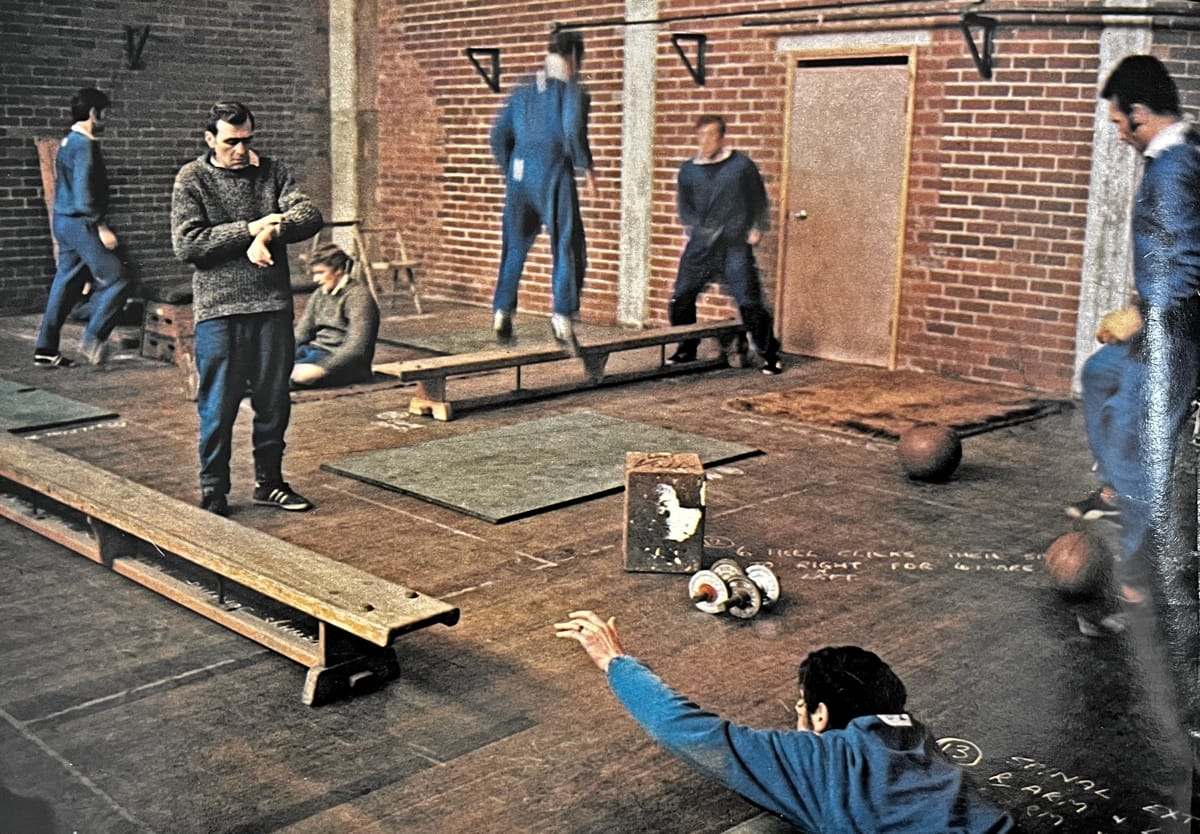 A photo of Elland Road's West Stand gym as described in the article, with bare brick walls, wooden benches and old weights, blurry players in various exercises (I can pick out Paul Reaney standing on the right next to I think Norman Hunter on the floor), with Les Cocker in the middle of them checking his watch